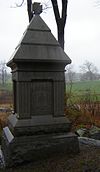 A granite monument with a pyramidal peak, capped by a maltese cross, etched with an inscription, "22nd Mass Infantry." In the background is a broad field and, in the far distance, a brick farmhouse.