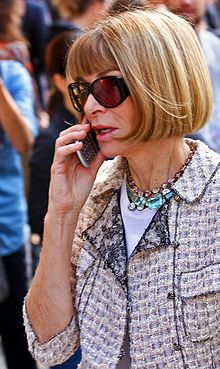 A woman with brownish hair, lit by the sun from outside the top right of the image, is seen from her front left. She is wearing a light-colored short-sleeved collared jacket with elaborate jewelry, a white top beneath it, and sunglasses. In her right arm she is holding a cell phone to her mouth; she is apparently in the midst of a conversation