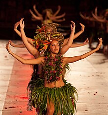 Danseuses tahitiennes sur scène, en train de danser en levant les bras les unes derrière les autres, vêtues de pagnes de feuilles et couronnes de fleurs