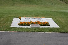 A grave marked by an angled slab of marble engraved with the names of Diefenbaker and his wife and surrounded by plantings of small marigolds, and an additional plaque in bronze. Beyond it a lawn stretches away.