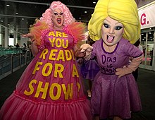 A photograph of two people; on the left is a female-presenting individual with pink hair wearing a pink dress with the text "Are you ready for a show?", and on the right is someone wearing a costume with blonde hair and a purpose dress with the text "Drag is magic".