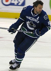 Hockey player in dark blue Vancouver uniform. He leans forward, holding his stick aloft.