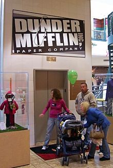 Трое взрослых и ребенок в коляске перед закрытыми дверями лифта. Над ними вывеска, на которой белыми буквами на черном фоне написано «Dunder Mifflin Paper Company».