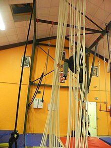 Photographie d’une personne suspendue à des cordes verticales.