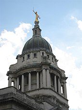 The stone dome at the top of a building, columns below, with a bronze statue on top of a woman with a sword in one hand and a pair of scales in the other