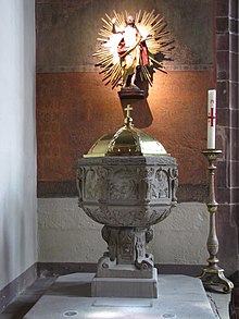 photographie en couleur montrant une cuve baptismale en grès gris coiffée d’un couvercle doré, avec à l’arrière-plan un mur peint d’une fresque rouge et décoré d’une statue dorée de Jésus