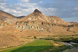 A conical mountain composed of many buff and pink rock layers rises above a small river bordered on one side by a green field. Black rock layers cap the mountain.