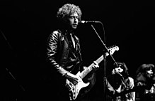 Dylan, onstage and with eyes closed, plays a chord on an electric guitar.