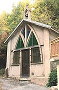 Photographie d'une chapelle en lisière de forêt, avec des vitraux verts et bleus ainsi que la porte marron en bois.