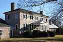 A photograph of a many-windowed stone building with a red-bricked chimney and a black roof surrounded by trees and green shrubs all under a clear, blue sky