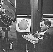 A young man sits holding a microphone in his left hand while manipulating the console of an apparatus with his right. To his left a large television camera is trained on a large, circular cathode ray tube display.