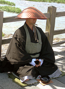 monk in brown robes with conical hat kneeling by alms bowl