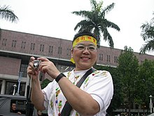 Josephine Ho hat kurzes schwarzes Haar und trägt ein gelbes Band im Haar, auf dem in roter Schrift etwas auf Chinesisch steht. Sie trägt eine Brille und lächelt in die Kamera. Sie steht im Freien und hält eine kleine Digitalkamera in den Händen.