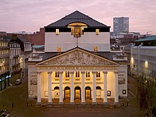 Photographie de nuit de la façade éclairée du théâtre : le péristyle aux huit colonnes surmontées du fronton sculpté précédant l'entrée du grand hall.