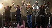 The members of Megadeth standing next to each other at a concert. They are holding their hands up.