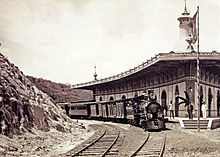 Photograph of a steam locomotive and train sitting on a curved section of track next to a curved platform and station building