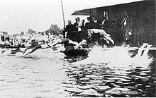 Plusieurs hommes en maillot en train de plonger dans l'eau depuis un ponton où se trouvent d'autres hommes en costume.