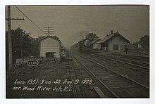 A steam locomotive pulls a train next to a small train station. Several tracks are visible.