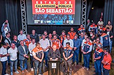 Num palco, há uma dezena de pessoas, com uma à frente, Luiz Inácio Lula da Silva, falando ao microfone. Ao fundo, há um cartaz com os dizeres: "Estado de calamidade pública: São Sebastião", em letras garrafais e com uma sirene de alerta ilustrada entre as palavras.