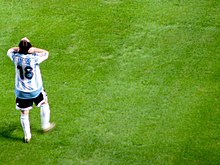 Photographie couleur. Homme revêtant la tenue de l’équipe d’Argentine de football se tient debout, de dos, sur une pelouse. Sur son maillot est marqué « Messi » et « 18 ».