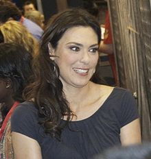 A Caucasian woman with brunette hair wears a dark grey blouse.