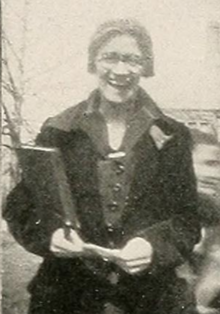 A young white woman, standing outside, smiling broadly, wearing a dark coat and glasses; she is holding a book and a slip of paper or cloth in her hands