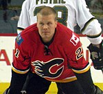 An ice hockey player stands facing the camera bent over at the waist, looking off into the distance. He is wearing a red jersey with black sleeves below the elbows. The jersey's crest is a stylized all black "C" made to look as if the curved part of the "C" has caught on fire. The shoulders have small patches on them, the player's left shoulder is the flag of Canada, on the players right is the flag of Alberta.