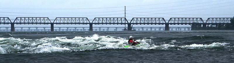 Kayakiste sur le fleuve Saint-Laurent à la vague devant Habitat 67. À l'arrière-plan, le pont Victoria. À l'horizon, le pont Champlain.