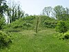 A large mound covered with grass and surrounded by brush and trees. A thin stairway ascends to the mound's summit.