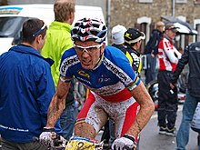 Homme sur un VTT en tenue bleu-blanc-rouge maculée de boue avec des spectateurs en arrière-plan.