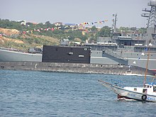 A partial view of a white yacht with Russian lettering, a partial view of a black submarine with lettering АЛРОСА and a partial view of a grew warship.