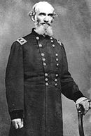 Black and white photo of a balding and gray-bearded man wearing a dark military uniform with the two stars of a major general on his shoulder tabs. He grips a chair in his left hand.