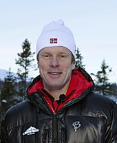 A man standing, facing the camera, smiling, wearing a cap.