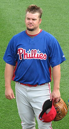 A fair-haired young man wearing a blue baseball jersey and white pinstriped pants stands on a grass field holding a baseball cap and mitt in his left hand