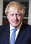 A close-up photograph of Boris Johnson, a smiling middle-aged man with ruffled-up blonde hair, a dark blue suit and light blue tie in front of a bookcase.