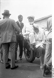 Caracciola sits on what appears to be the back of a vehicle. A young woman sits in front of him.