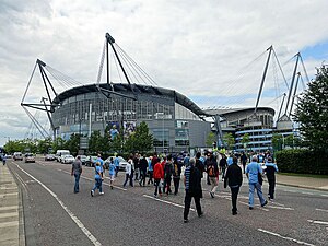 A grey stadium exterior with glass fronting. Adjoining it is a spiral walkway made of concrete, rising almost to the full height of the structure.