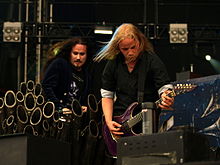 Image d'un homme blond jouant de la guitare devant un homme aux longs cheveux bruns devant des tuyaux d'orgue.