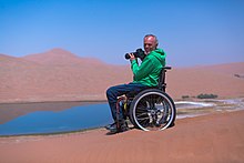 Ein Mann mit Kamera in der Hand sitzt in einem Rollstuhl, der auf einer Sanddüne steht, und blickt lächelnd zum Fotografen; im Hintergrund ist ein von Wüste umgebener See zu sehen, in dem sich der klare blaue Himmel spiegelt.