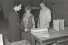 Monochrome photograph of William Waldegrave's visit to the heat laboratory at the University of Salford in 1981. Waldegrave is shown stood with Stuckes, the then head of the laboratory, and John Ashworth. They are photographed behind a table holding anechoic poly blocks.