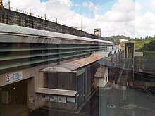 Photo du barrage de Petit-Saut en Guyane vu de l'intérieur d'un bâtiment, sur laquelle figure le mur du barrage et des bâtiments