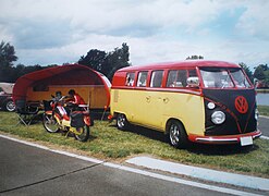 Un VW Combi Split Samba accompagné de sa caravane et d'un cyclomoteur, tous les deux assortis. La face avant du Combi est couverte d'une housse pour protéger sa peinture des gravillons de la route.