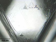 Треугольная грань кристалла с треугольными ямками травления, самая большая из которых имеет длину основания около 0,2 мм.