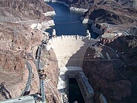 Aerial view of a white structure located between mountains with a lake, roads in the left, power lines in the right and a pylon with some wires in the low part