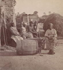 An African American family, photographed by O'Pierre Havens, circa 1868 Little Bo Peep. Group of men and women seated outside, child peeking out of a barrel in the foreground.png