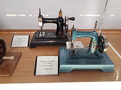 Deux machines à coudre de taille réduites sont exposées dans une vitrine. Celle de devant est peinte en bleu-gris, celle de derrière est noire. Toutes les deux sont conçues pour être actionnées à la main.