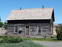 Photograph of a log cabin in a manicured setting with agricultural fields and buildings in the far background