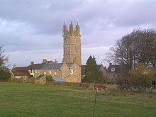 Yellow stone church tower above other buildings of the same stone. In the foreground is a grassy field with cows