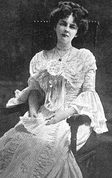 A white woman, seated in a large chair; she has dark hair in a bouffant updo, and is wearing a light-colored lacy corseted dress with elaborate details; her hands hold a fan in her lap