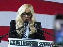 A woman with blonde hair speaking at a podium into several microphones. She wears large glasses. The background is a series of red and white horizontal stripes.